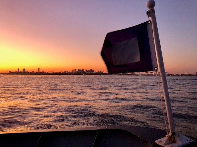 This image captures a serene sunset over a body of water, with a city skyline visible in the distance. A flag on a boat in the foreground adds a focal point to the scene. The sky displays a beautiful gradient of colors, from warm oranges and yellows near the horizon to cooler purples and blues above. The calm water reflects the sky's colors, creating a peaceful and picturesque view.